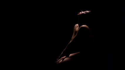 Naked young woman sitting in darkness with her arms on knee and looking up, panorama with copy space