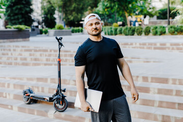 Handsome man holding laptop in his hand standing near electric scooter. Active lifestyle, backpacker traveler.