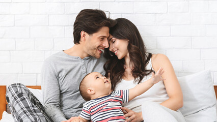 A young couple is sitting on a bed in their bedroom, with their baby sitting on the mothers lap. The couple is smiling at each other while the baby is looking up at them.