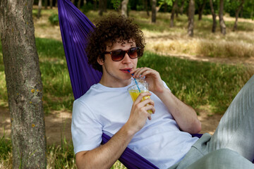 Obraz premium A young guy is lying in a purple hammock drinking orange juice. A young guy with curly hair is relaxing on a hammock on a hot summer day.