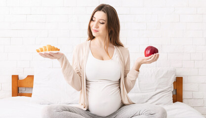 Healthy nutrition and pregnancy. Pregnant woman choosing between croissant and apple, sitting on bed at home