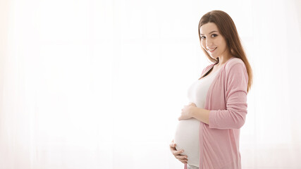 A pregnant woman is smiling while gently holding her belly. She appears joyful and content as she...