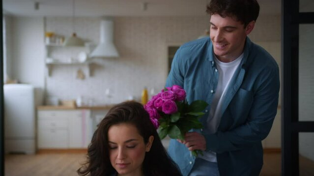 Loving Caucasian man guy male romantic husband boyfriend come living room make surprise present bouquet flowers roses to woman girl female wife girlfriend using mobile phone valentines day anniversary
