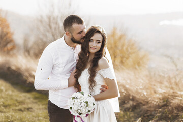 A bride and groom are hugging each other in a field. The bride is holding a bouquet of flowers