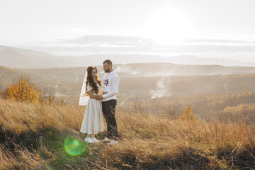 A couple standing on a grassy hillside, with the man wearing a wedding ring. The woman is wearing a white dress and a veil. The scene is peaceful and romantic