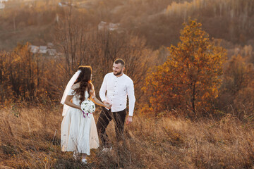 A bride and groom are walking through a field of tall grass. The bride is holding a bouquet and the groom is holding her hand. The scene is peaceful and romantic