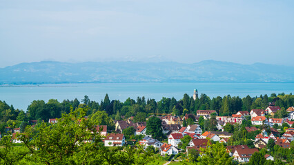 Germany, Bad schachen tower, houses, buildings, and green trees surrounded by silent bodensee lake water early in the morning after sunrise, aerial panorama landscape view to swiss mountains