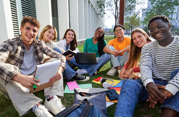 Portrait of a multiethnic group of student friends outside university campus looking at camera smiling, sitting on the grass gathered to study together.