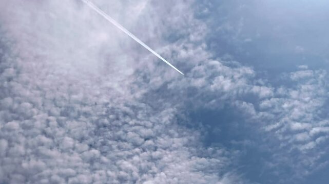 An airplane with jet contrail crossing the sky under altocumulus clouds covering the blue sky. Cloudscape.