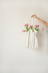 Delicate hand holds a white cloth bag brimming with fresh pink tulips against a light background. Minimalist and elegant floral arrangement. Spring and summer vibes in soft pastel tones