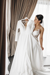 Fototapeta premium A woman is standing in front of a white dress hanging on a hanger. She is wearing a veil and she is getting ready for a wedding