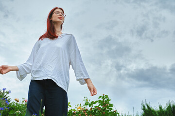 Young red-haired woman in white shirt and glasses standing in flower field under cloudy sky. Outdoor photography of nature and leisure.