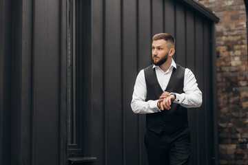 A man in a black vest and white shirt is standing in front of a brick wall. He is wearing a watch and he is checking the time