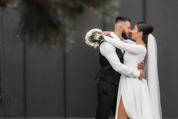 A bride and groom are embracing each other in front of a black wall. The bride is wearing a white...