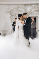 Naklejka premium A bride and groom are dancing in a foggy room. The bride is wearing a white dress and the groom is wearing a black suit