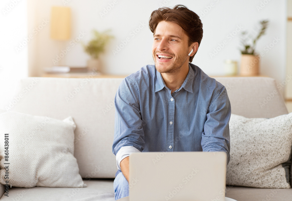 Poster a man sits on a couch, laughing while using a laptop. he is wearing a blue button-down shirt and ear