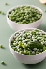 Two white bowls filled with frozen green peas, with a fresh basil leaf on a green background. 