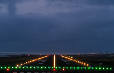Runway illuminated with green and yellow lights at night with a view of the ocean in the background. The sky is dark and cloudy