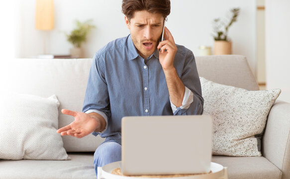 A frustrated man sits on a couch, looking at his laptop screen while talking on his phone. He is likely having technical difficulties.