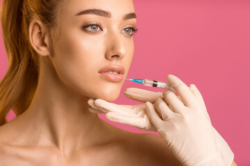 A young woman with long red hair is receiving lip injections from a medical professional wearing white gloves. The woman is looking to the side and the gloved hand holding the syringe
