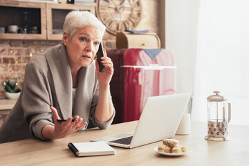 A senior woman with white hair sits at a kitchen table talking on a mobile phone while gesturing with her other hand. She is sitting in front of a laptop computer and a notebook.