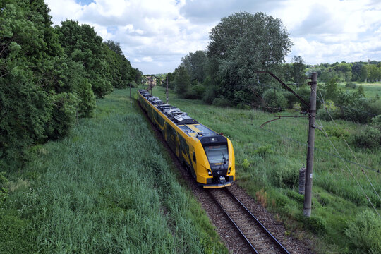 A modern yellow and black electric passenger train heads out of Tukums through a lush green landscape. Tukums, Latvia 06 04 2024 