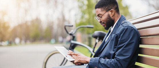 A professional black man is seated on a park bench, using a tablet with his bicycle next to him. The scene is peaceful and modern, highlighting a balance of technology and relaxation.