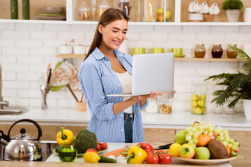 A woman smiles while looking at a laptop in her kitchen, surrounded by fresh produce.