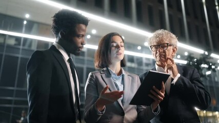 Senior boss talking to workers outside at night. Men and woman communicating near modern business center. Male and female in official clothes having active discussion outdoor. Ethnic diversity.