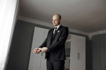 A man in a suit is standing in front of a window, adjusting his tie. Concept of formality and professionalism, as the man is dressed in a suit and tie. The window provides natural light