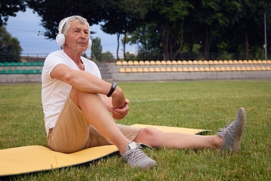 Athletic sporty Caucasian senior man having break while training at stadium sitting on yoga mat listening to music enjoying rest during outdoor workout