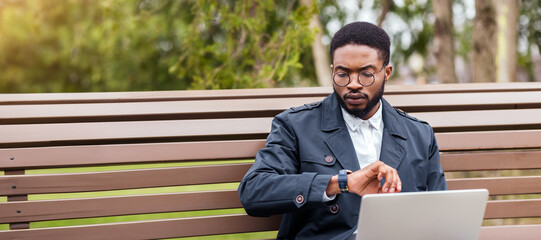 A professional black man uses his laptop on a park bench, checking time. The scene is tranquil and modern, emphasizing outdoor work-life balance.