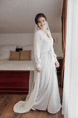 A woman in a white dress stands in front of a bed. She is smiling and she is happy