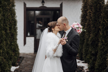A bride and groom are kissing in front of a building. The bride is wearing a fur stole and the groom is wearing a suit