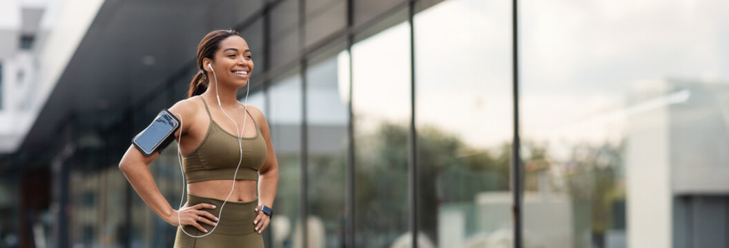 Attractive young black woman in sports outfit listening to music in earphones, using mobile device during jogging on city street, empty space. Urban sports, outdoor fitness concept - Powered by Adobe
