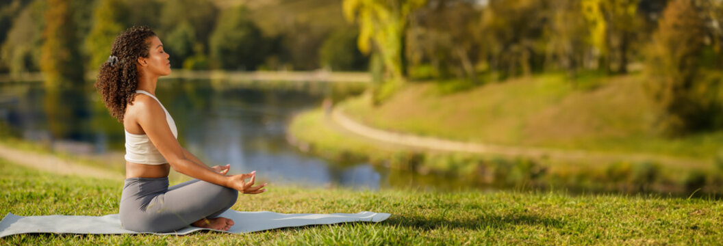 Side view of millennial woman in sports outfit meditating on yoga mat at lake in green park outside. Lady practicing breathing exercises in nature for inner peace and harmony outdoor, copy space
