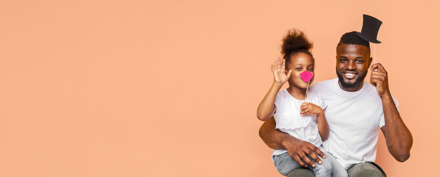 A Black father and his daughter are wearing party props while posing in front of a peach-colored backdrop. The father is wearing a black top hat, and the daughter has a pink mustache, copy space