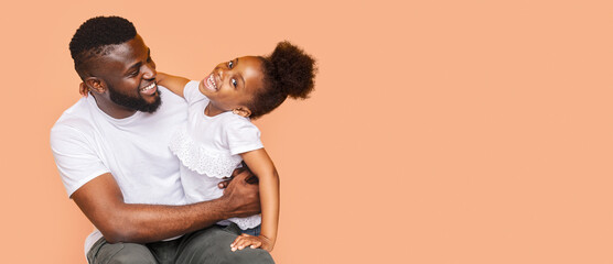 A black father and his little daughter are sitting together in a studio, laughing and enjoying each others company. The background is a soft orange color, copy space