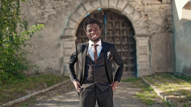 Happy African American groom standing in front of entrance to old castle or church and positively smiling at camera. Handsome man with elegant black suit waiting to start wedding ceremony outside.