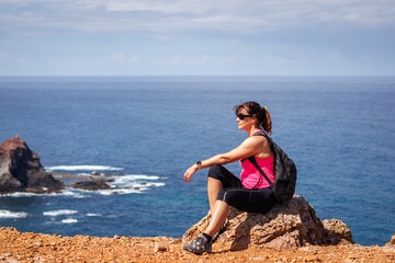 Sporty mature woman resting during hiking in Algarve seashore. Hiker is enjoying view at Atlantic ocean from cliff in Portugal