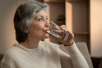 Fototapeta premium An elderly woman is sitting on the couch in her house in the evening and drinking a glass of water. She is wearing a beige sweater and is sitting in a relaxed position.