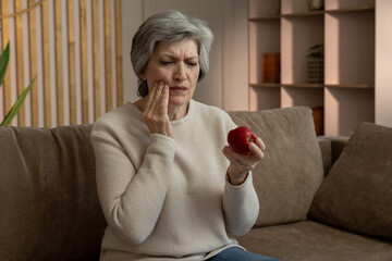 A woman sits on a couch in her living room and take a bite of an apple. She grimaces in pain,...