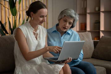 A daughter patiently teaches her mother how to use a laptop in their living room. The mother is focused on the screen and appears to be learning new skills