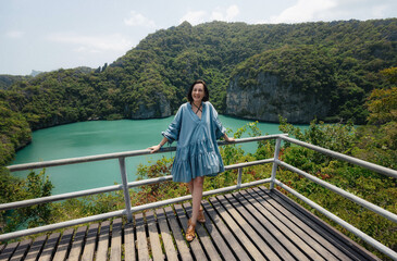 Fototapeta premium A woman wearing a blue dress leans on a railing and enjoys a view of a scenic lagoon surrounded by lush green hills on a sunny day