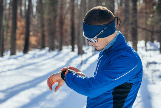 Cross-country skier in blue jacket checks his smartwatch while standing in snowy forest, enjoying winter day. Man athlete looks at workout indicators on smart watch, heart rate and pace tracking. - Powered by Adobe
