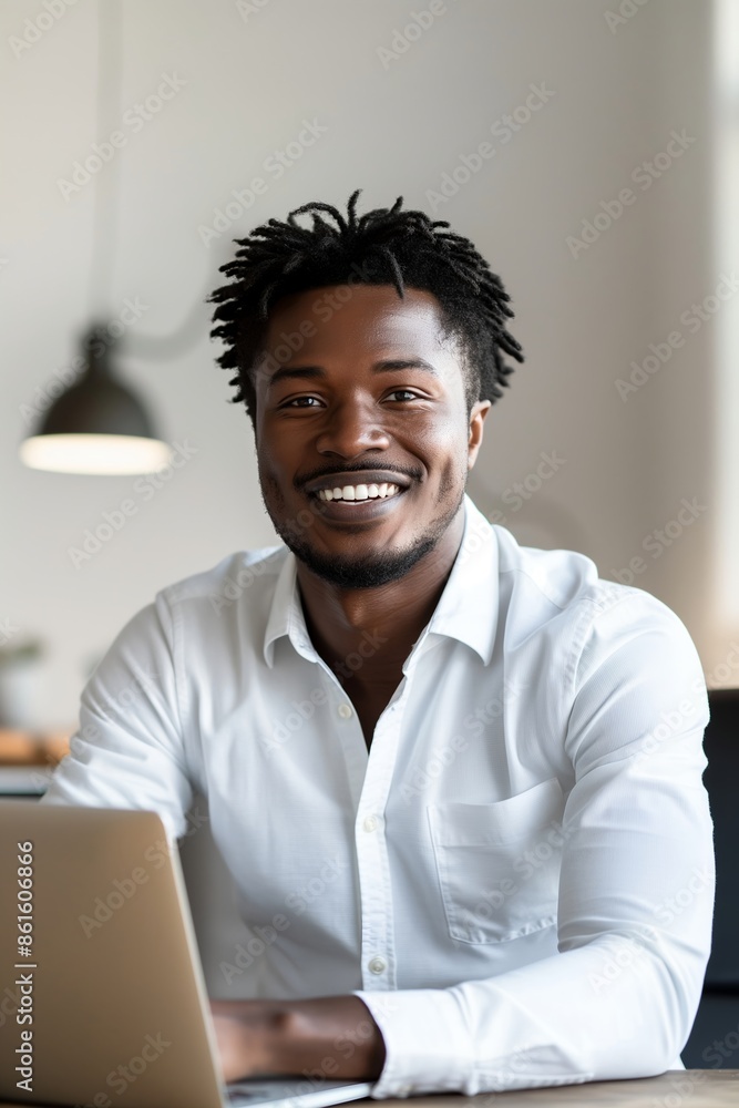 Wall mural Attractive African American businessman sitting at his desk with a laptop, smiling and looking at the camera, wearing a white shirt, with short black hair and beard, against a light background.