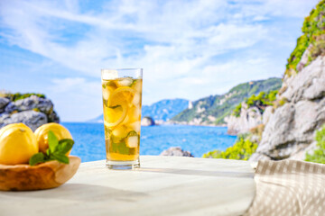 Image of copy space for drinks and food and products. Glass of mineral water with lemon slices, ice and straws on white table with blurred seascape and rocky mountains background.