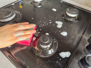 female hand in gloves with rag sponge a foam cleaning gas stove at home kitchen