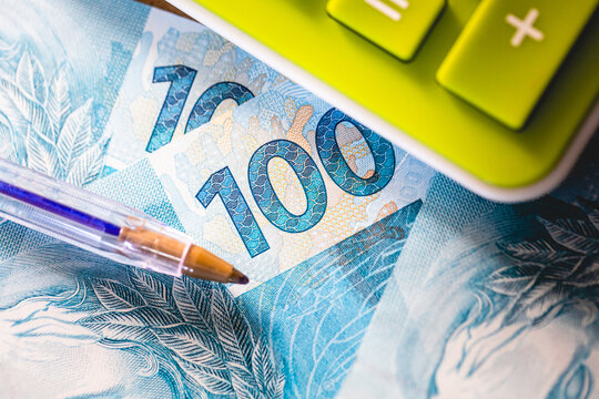 Vista superior em closeup de notas de 100 reais com uma calculadora e uma caneta na composi&ccedil;&atilde;o. Real BRL - moeda do Brasil. Impostos, imposto de renda, fundo de garantia -  fgts e sal&aacute;rio m&iacute;nimo.