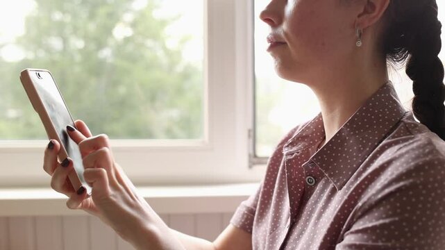 Side view of woman's presses quickly her fingers on smartphone screen, earning virtual crypto coins. Taping for online earning of tokens. Crypto app and farming. Window in background